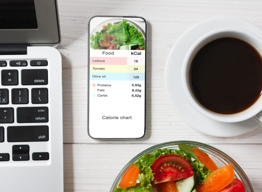 Calorie Counter Concept. Modern Smartphone With Opened Application For Counting Calories On Screen Flat Lay On Table With Coffee, Laptop And Vegetable Salad On White Table