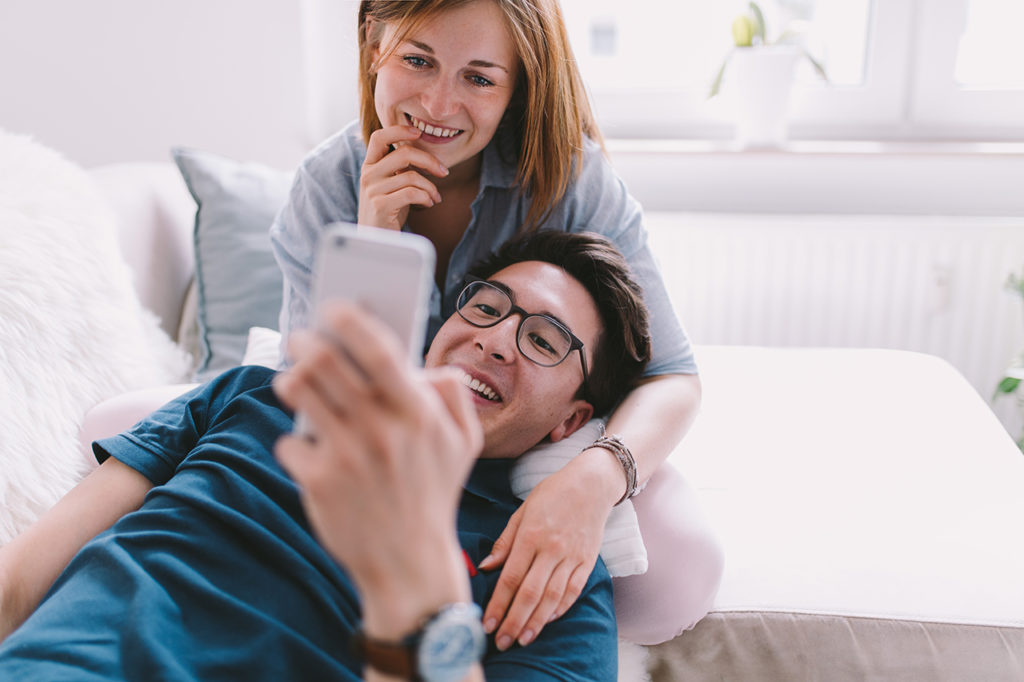 Happy relationship image of a couple. CoupleSmartphone Smiling Casual Wear Glasses Indoor Bright Room Cozy Togetherness Relaxation This image shows a young couple enjoying a cozy moment together. The man is lying back on a white cushion, holding a smartphone in his hands, with a woman sitting behind him. She has her chin resting on his shoulder and is smiling as she looks at the smartphone screen. Both individuals are casually dressed; the man is wearing glasses and a blue T-shirt, and the woman is in a light blue shirt. They appear to be in a bright, sunlit room with a window in the background, suggesting a relaxed, domestic setting.