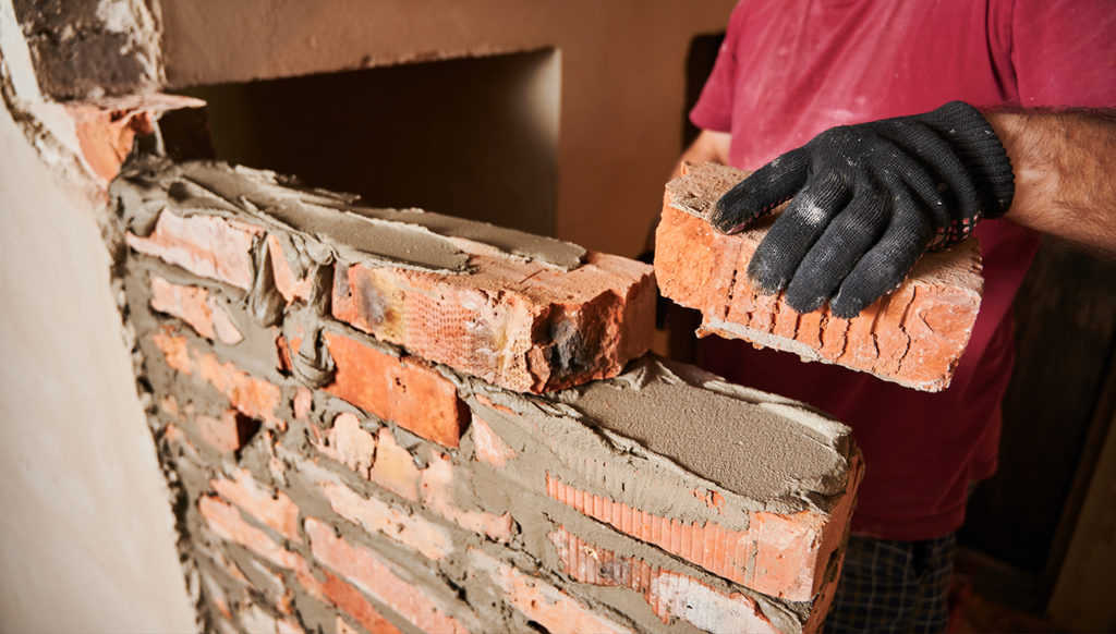 Visual metaphor for rebuilding after a breakup or divorce: Close up of male hand in work glove laying brickwork in building under construction. Man builder placing brick on block course structure with cement mortar.